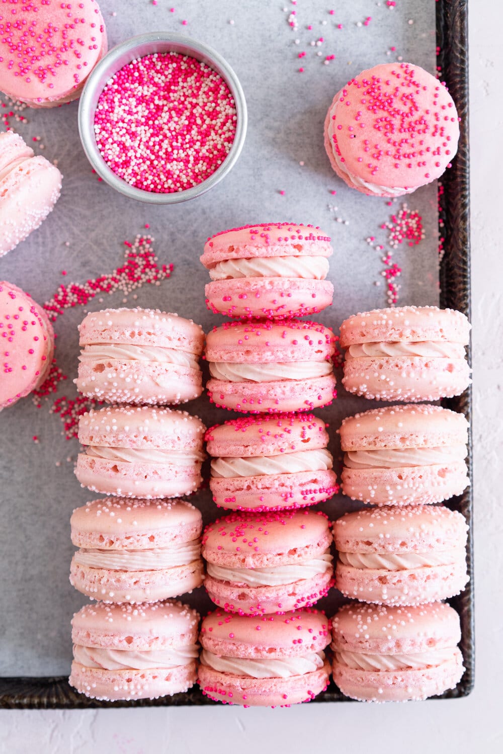 filled pink macarons on a sheet pan with a dish of pink sprinkles.