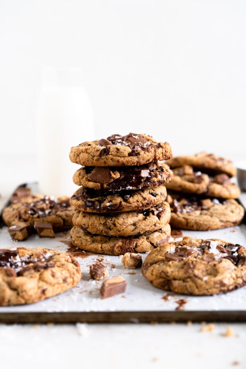 Stacked up chocolate chip cookies with extra cookies in the foreground and a bottle of milk in the background